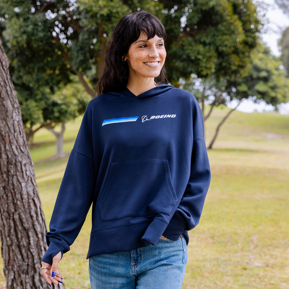 A woman in a Boeing Women's Signature Stripe Hoodie by BOEING and jeans stands smiling in a grassy park with trees behind her.