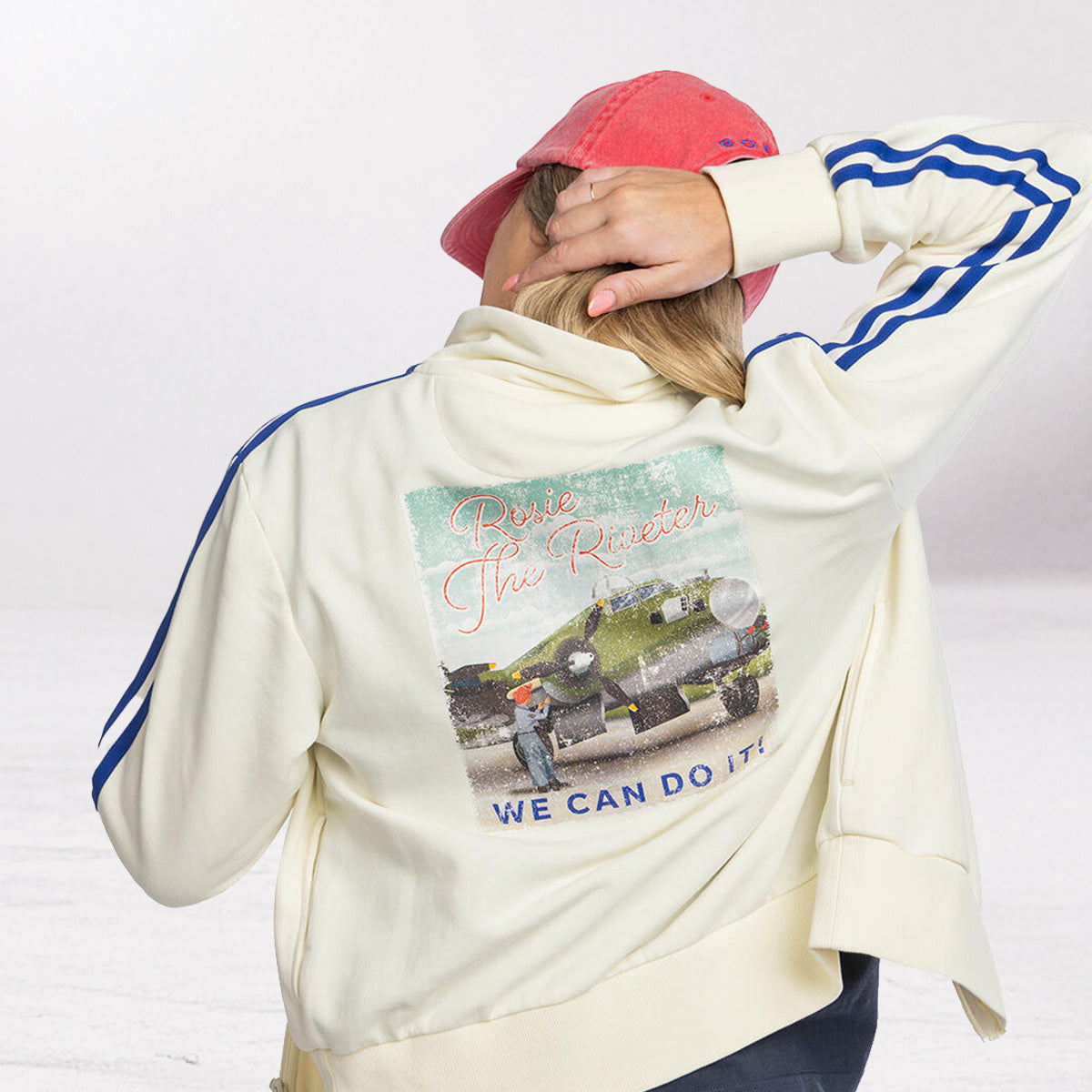 Person wearing a cream-colored Rosie the Riveter track jacket with a graphic design on the back, standing against a white background.