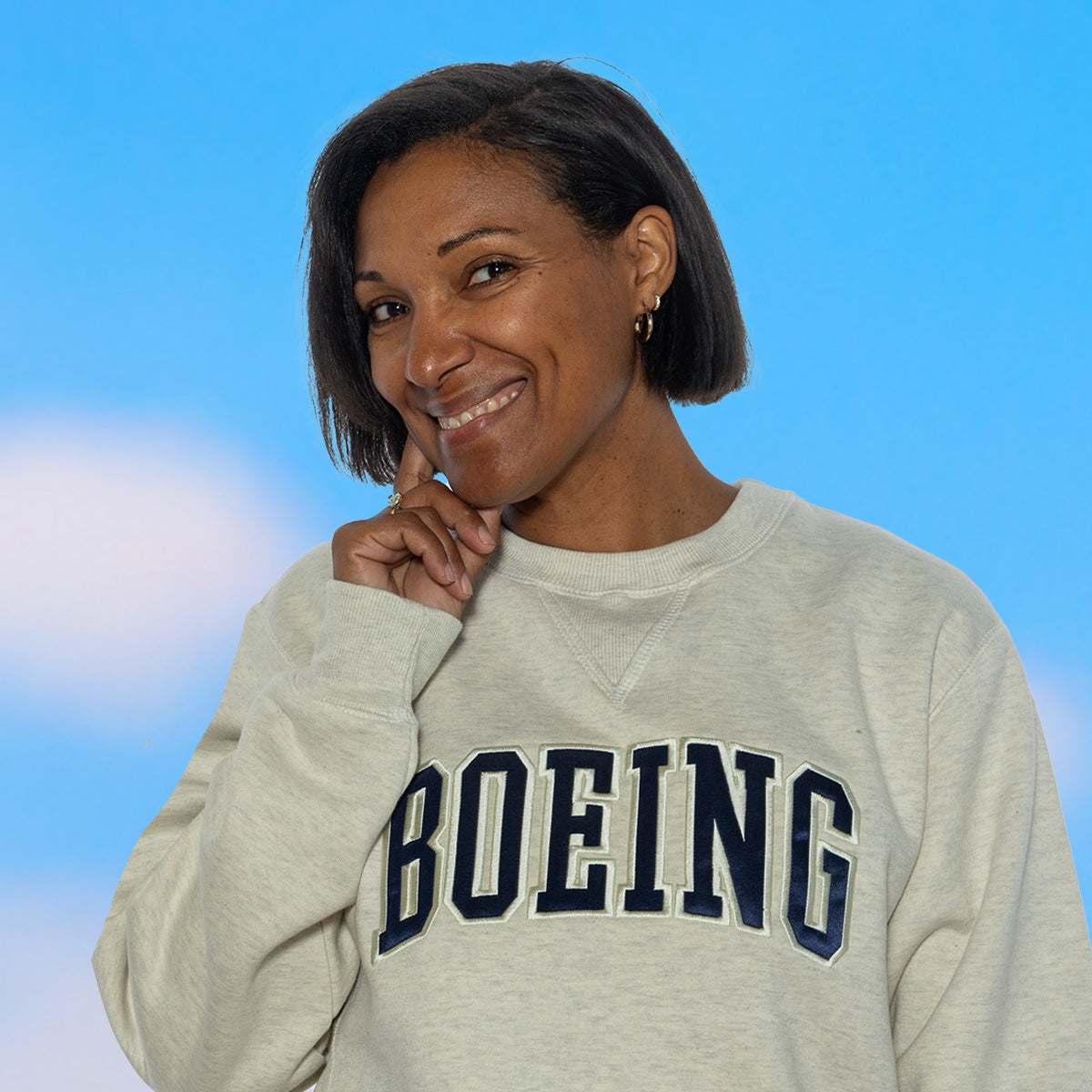 A person wearing a gray sweatshirt with Boeing's varsity logo printed prominently on the front, set against a blue sky background.