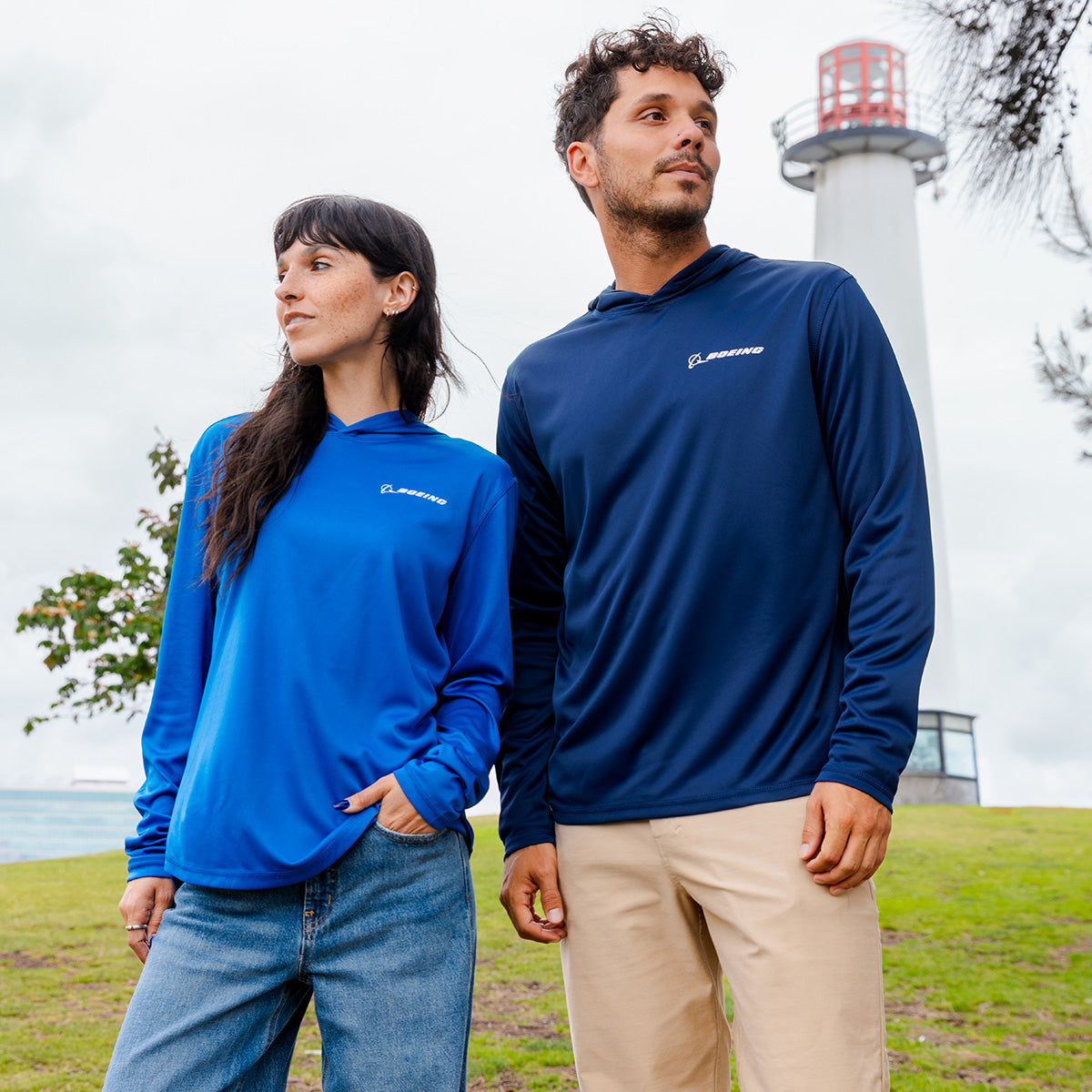 A woman in a bright blue UPF 50+ shirt and jeans and a man in the BOEING Signature Logo Unisex UV Hoodie stand outdoors near a white lighthouse, looking off into the distance.