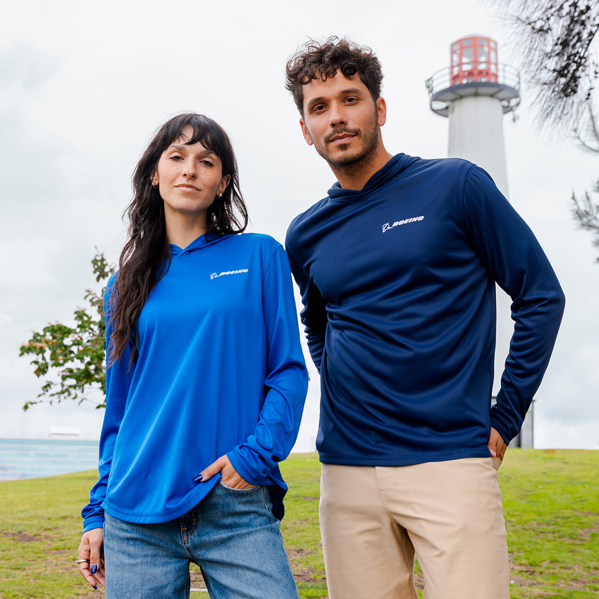 Two people stand outdoors near a lighthouse, each wearing a Boeing Signature Logo Unisex UV Hoodie by BOEING. One pairs it with light jeans, the other with khaki pants. The sky is cloudy, with grass and a tree in the background.