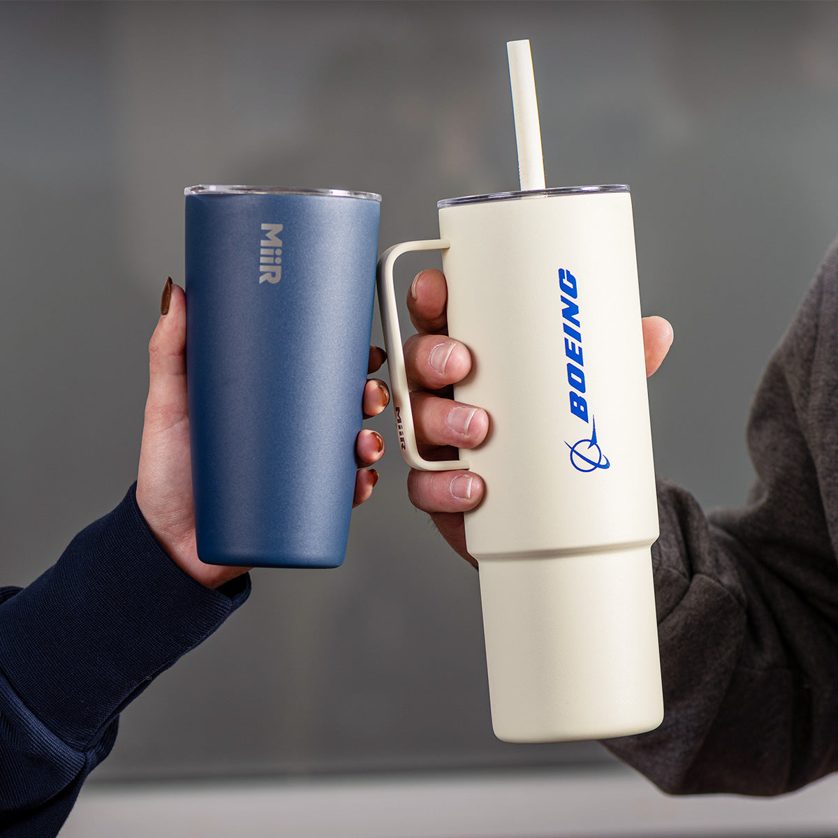 Close-up of two people holding MiiR x Boeing branded drinkware that features both logos. One is a blue tumbler with white logos and the other is a white bottle with silicone straw and blue logos.