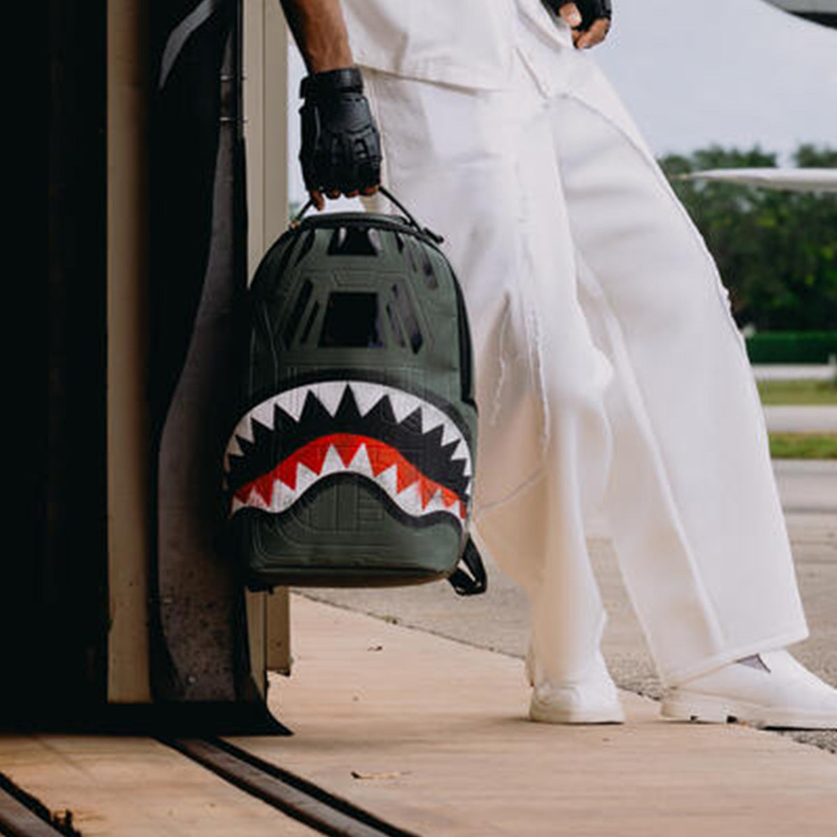 A person in white pants and shoes leans against a wall, holding the BOEING Sprayground Boeing AH-64 Apache Sharkmouth DLXSV Backpack. The focus is on the bag, which features tactical styling and a bold shark mouth design, with attention on the lower half of the body.