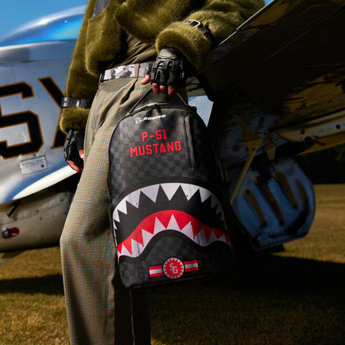 A person in a green jacket and checkered pants stands beside a silver plane, holding the BOEING Sprayground P-51 Mustang Sharkmouth DLXSV Backpack. Inspired by vintage fighter planes, this aviation-style backpack features bold shark mouth nose art and striking P-51 Mustang design details.