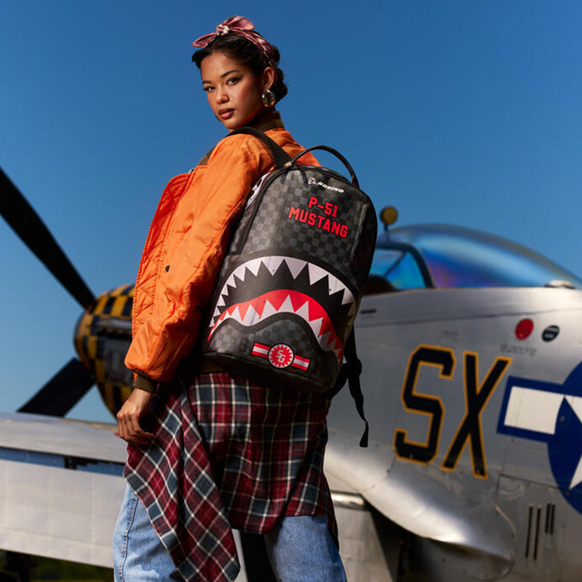 A woman stands outdoors in front of a silver airplane under a clear blue sky, wearing an orange jacket, plaid shirt, and headband. She holds the Sprayground Boeing P-51 Mustang Sharkmouth DLXSV Backpack by BOEING, which showcases aviation-inspired shark teeth graphics and design elements reminiscent of the iconic P-51 Mustang.