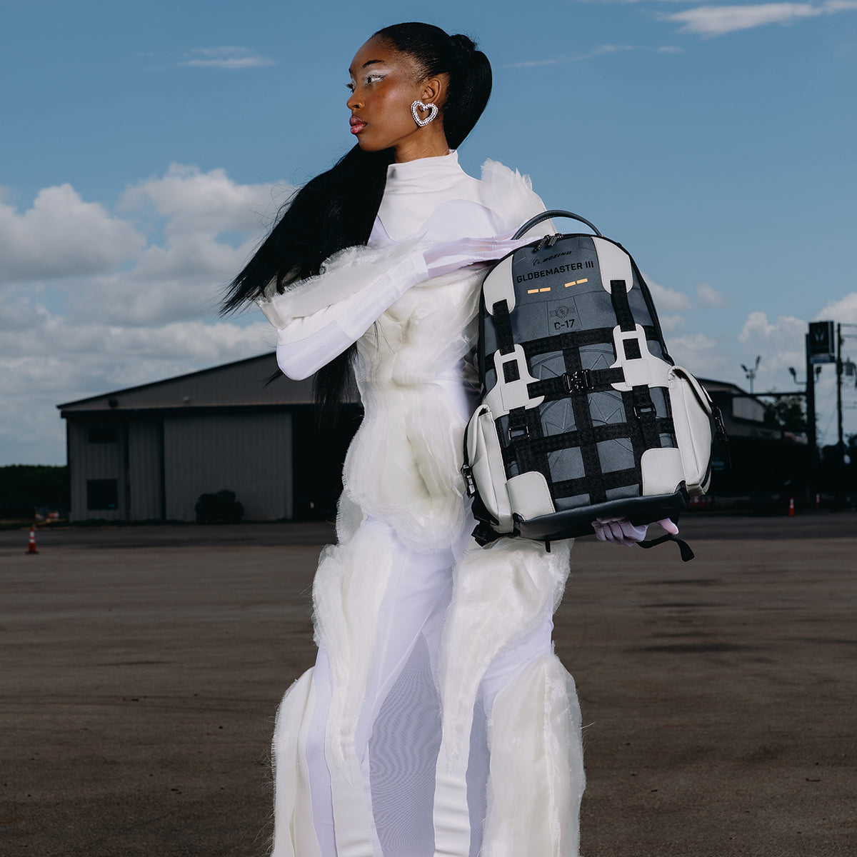A woman in a white, futuristic gown stands outdoors holding the BOEING Sprayground C-17 Globemaster DLXSV Backpack. She gazes to the side, with a blue sky and industrial buildings in the background.