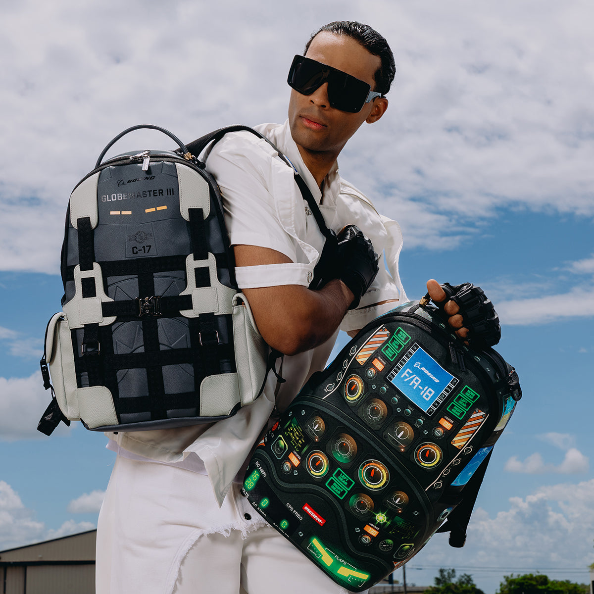 A person in white clothing and dark sunglasses poses outdoors under a blue, partly cloudy sky, holding two colorful, futuristic backpacks—one gray and black BOEING backpack with a dedicated laptop compartment, and the other mostly black with electronic-style designs and buttons reminiscent of the Sprayground Boeing F/A-18 Super Hornet DLXSR Backpack.