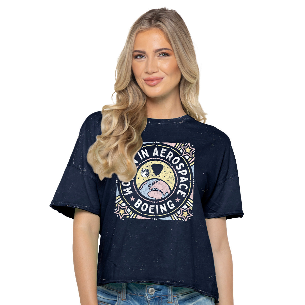 A woman with long blonde hair models the Boeing Women In Aerospace Space Womens T-Shirt by Boeing, a vintage-style navy blue tee in 100% cotton jersey featuring a colorful space-themed graphic, paired with blue jeans against a white background.