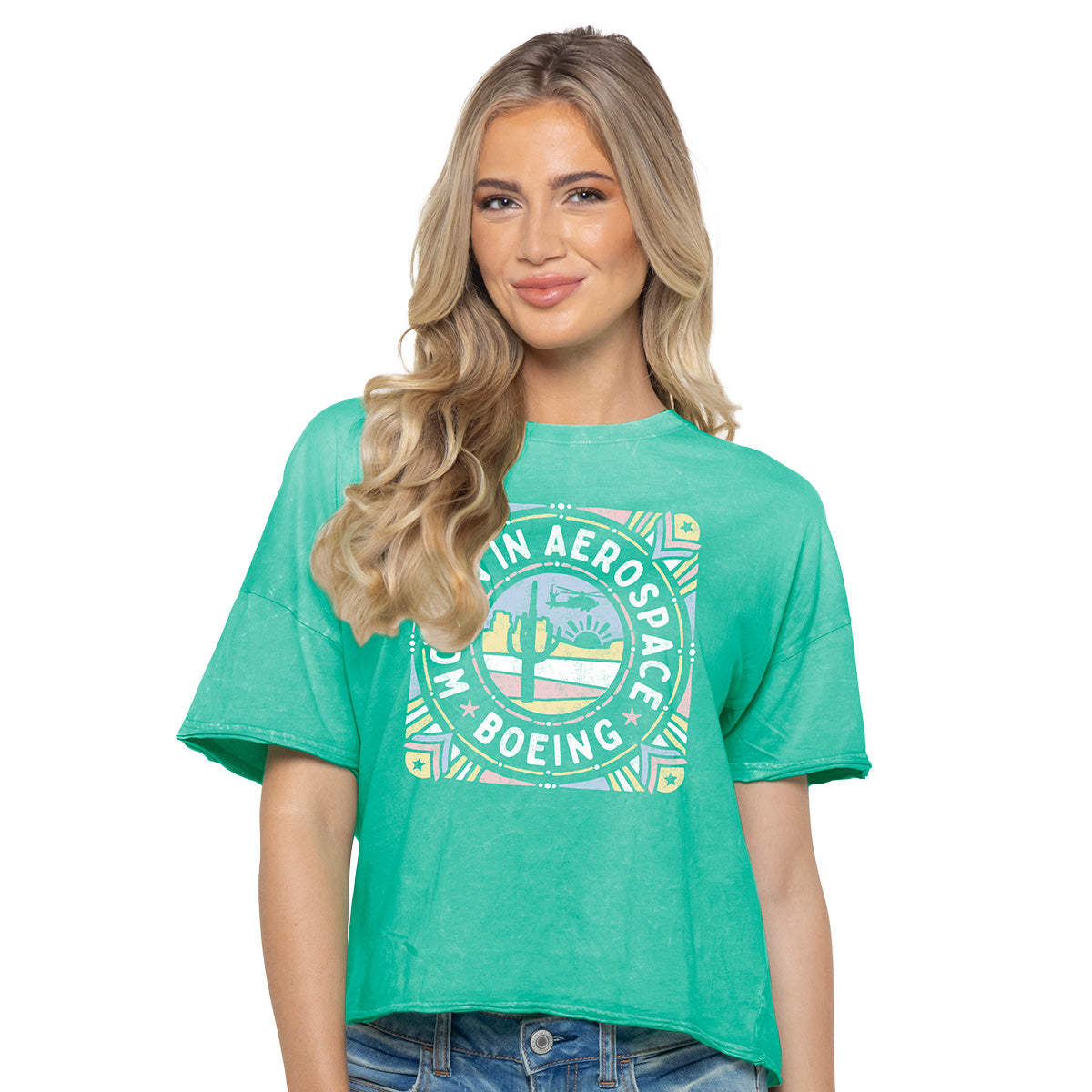 A woman with long blonde hair wears the Boeing Women In Aerospace Defense Womens T-Shirt, featuring a white circular Boeing logo, and light blue jeans, smiling in front of a plain white background.