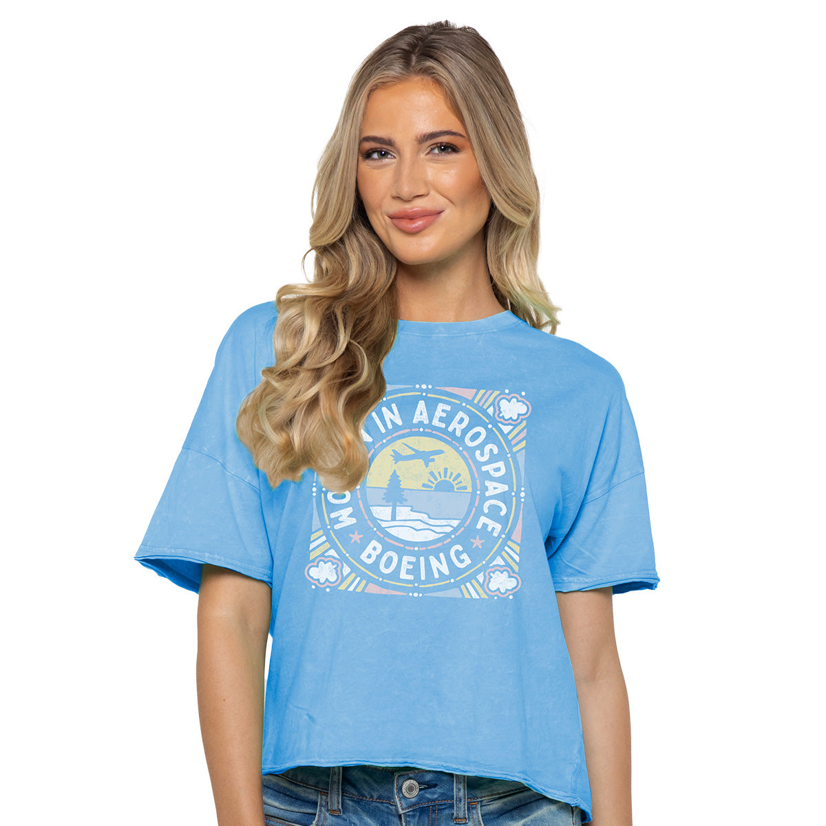A smiling woman with long blonde hair wears the Boeing Women In Aerospace Commercial Womens T-Shirt, featuring a round Boeing logo and graphic on soft, vintage-feel fabric. She stands against a plain white background.