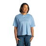 A woman wearing a light blue BOEING Women's Boardwalk T-Shirt in a relaxed fit with blue jeans stands against a white background, smiling and looking slightly to her left.