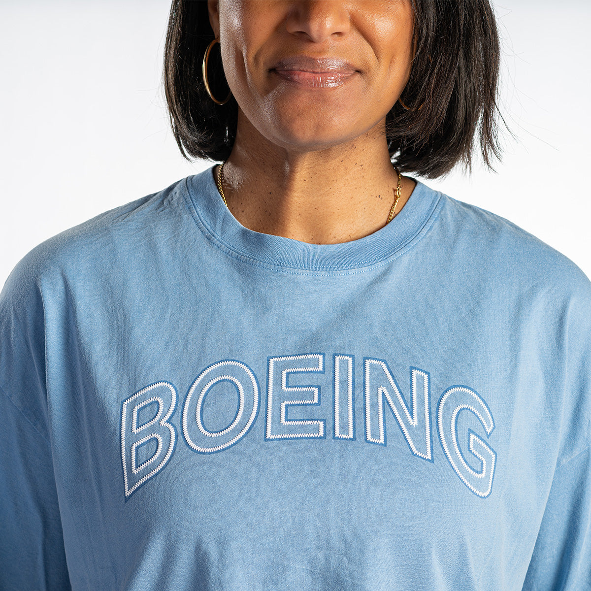 A woman with shoulder-length dark hair wears a light blue BOEING Women's Boardwalk T-Shirt in a relaxed fit, made from soft cotton jersey. Only the lower half of her face and upper torso are shown.