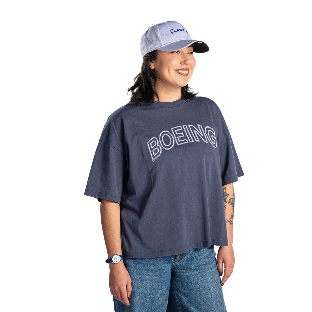 A person wearing a gray Boeing Women's Boardwalk T-Shirt by BOEING, a white and blue baseball cap, blue jeans, and a watch stands smiling against a white background.