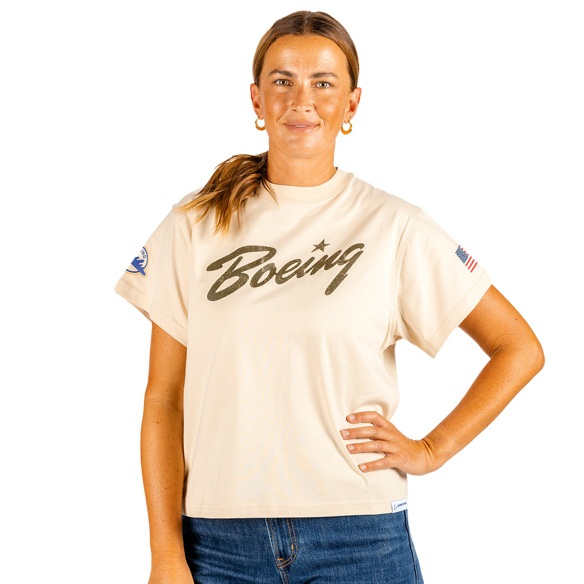 A woman with brown hair in a ponytail, dressed in blue jeans and the Boeing Heritage Script B-17 Flying Fortress Women’s T-Shirt by Boeing, stands with one hand on her hip. This beige tee celebrates aviation heritage, showcasing an American flag patch on one sleeve and a special patch honoring the B-17 Flying Fortress on the other.