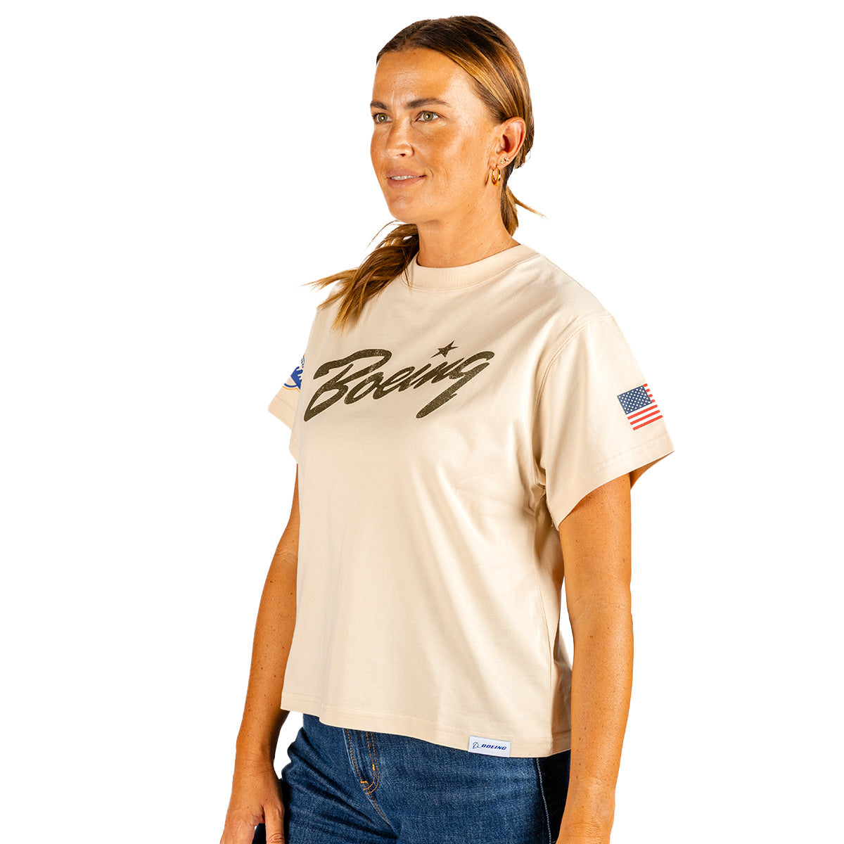 A woman stands against a white background wearing the Boeing Heritage Script B-17 Flying Fortress Women’s T-Shirt—a beige tee from Boeing featuring the brand’s bold script logo on the front and an American flag patch on the sleeve, celebrating classic aviation heritage.