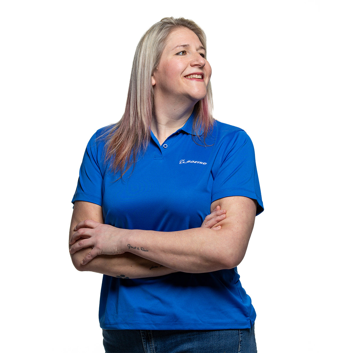 A person with long light hair stands against a white background, wearing The Boeing Stores moisture-wicking Boeing Signature Logo Womens Mesh Tech Polo in blue with a rib-knit collar, arms crossed, and smiling while looking to the side.