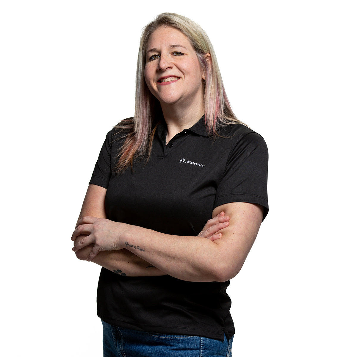 A person with long blonde hair stands confidently, arms crossed, wearing a sleek Boeing Signature Logo Womens Mesh Tech Polo by The Boeing Store and blue jeans. Their poised demeanor is highlighted against a pristine white background.