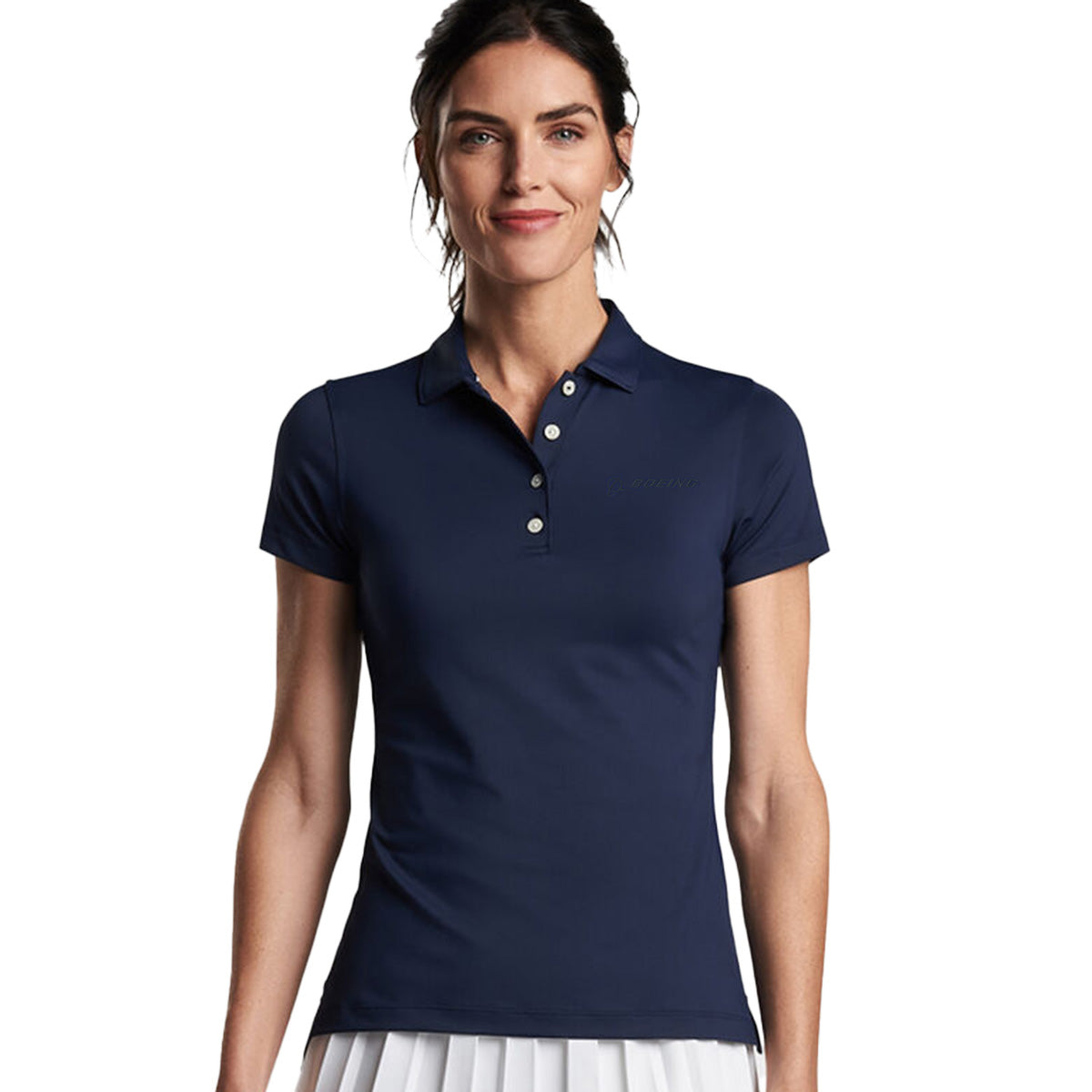 A woman with dark hair tied back is wearing the Peter Millar Boeing Women’s Button Polo in navy by Boeing and a white pleated skirt, standing against a plain white background and smiling slightly.