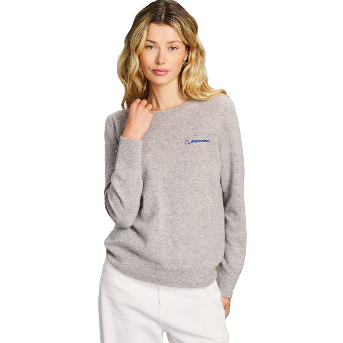 A woman with long blonde hair is wearing the Boeing Signature Women’s Cashmere Sweater by BOEING in light gray, which features a small blue Boeing logo on the chest, paired with white pants and standing against a white background.