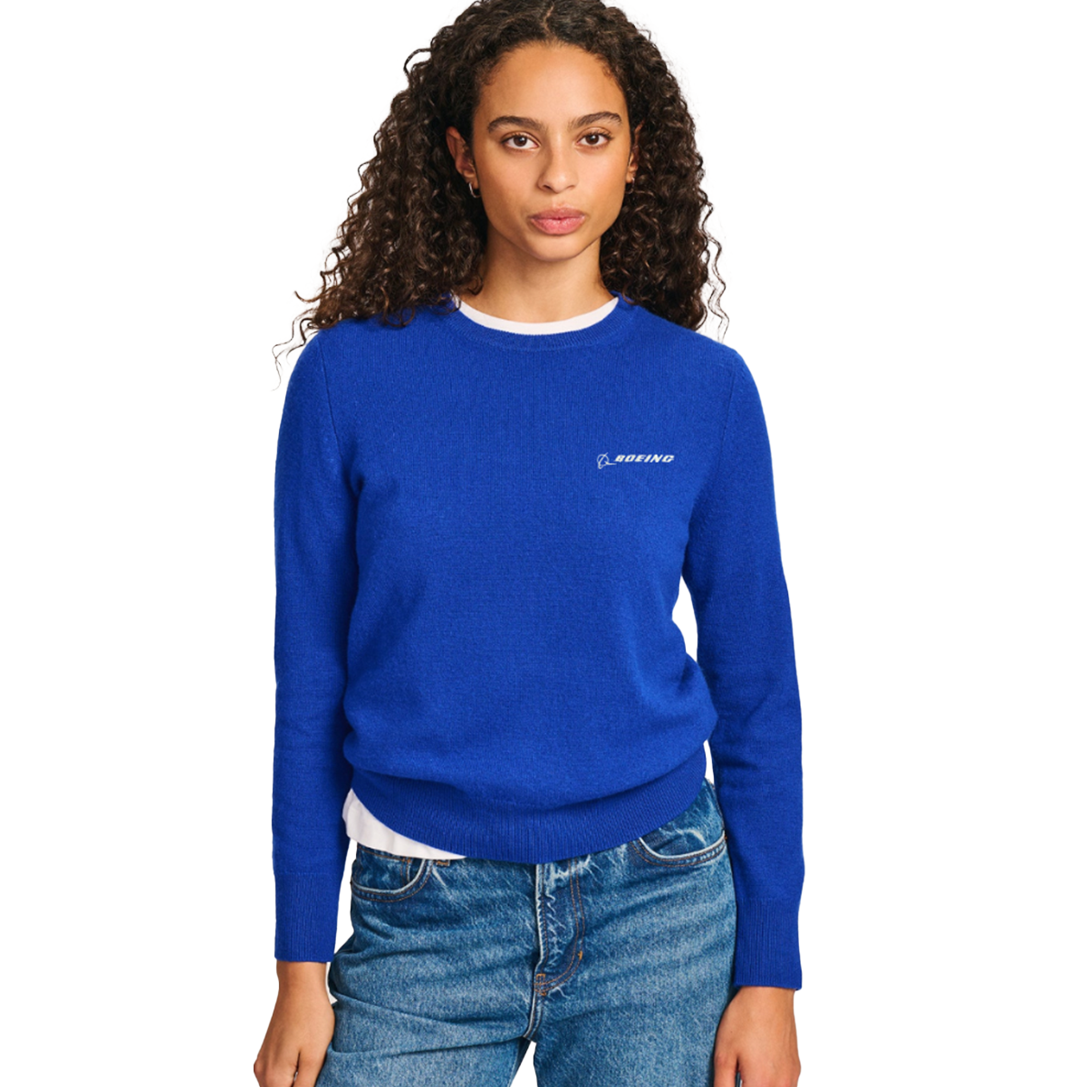 A woman with curly hair is wearing the Boeing Signature Women’s Cashmere Sweater in bright blue by BOEING, featuring “BOEING” on the chest. She has layered it over a white shirt and paired it with blue jeans, standing against a plain white background.