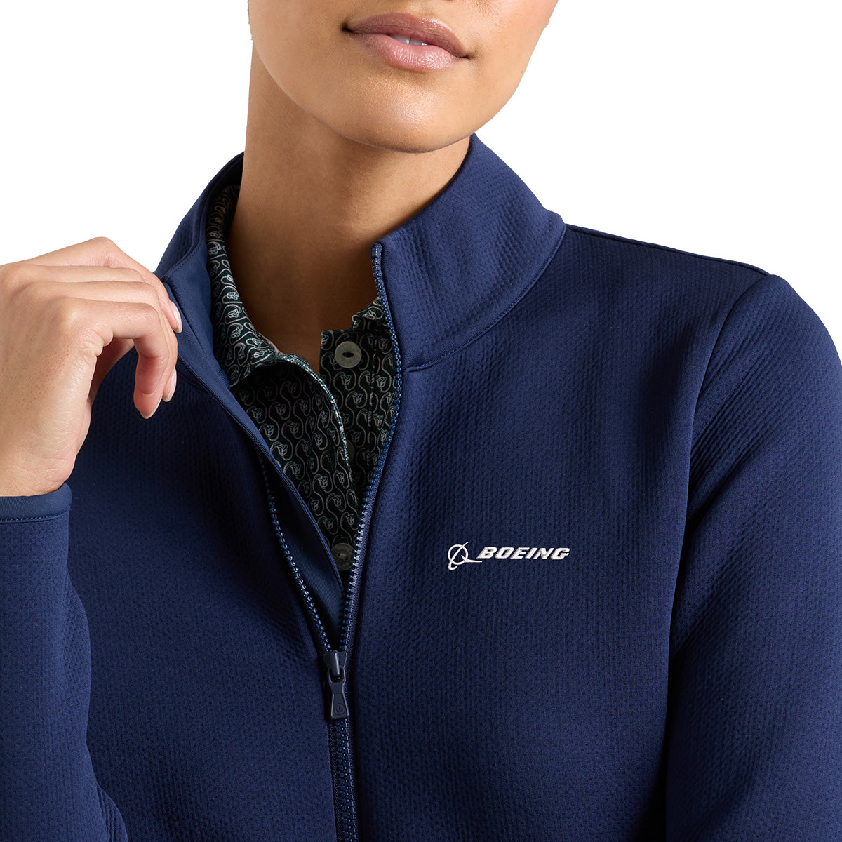 A woman wearing the Peter Millar Boeing Women’s Lumi-Knit Full-Zip jacket in navy blue, featuring the Boeing logo on the chest and four-way stretch fabric, layered over a patterned shirt. She is shown from the shoulders up against a white background.