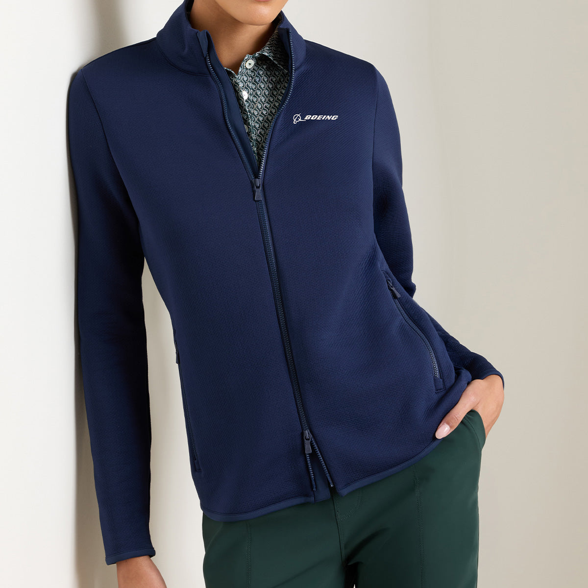 Wearing a navy blue Peter Millar Boeing Women’s Lumi-Knit Full-Zip jacket, featuring the Boeing logo on the chest and four-way stretch, layered over a patterned collared shirt and paired with dark green pants, this person stands against a neutral wall.
