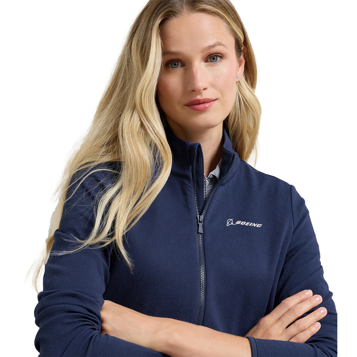 A woman with long blonde hair stands against a white background, arms crossed, wearing the BOEING Peter Millar Women’s Lumi-Knit Full-Zip in navy. This stylish jacket features a white Boeing logo and is crafted with four-way stretch for enhanced comfort and flexibility.