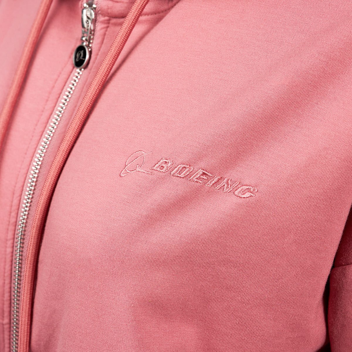 A close-up shows a person wearing the Boeing Signature Logo Womens Short Waist Hoodie by The Boeing Store. It features a pink color with an embroidered Boeing logo on the chest and has a silver YKK zipper with a stylish round black pull.