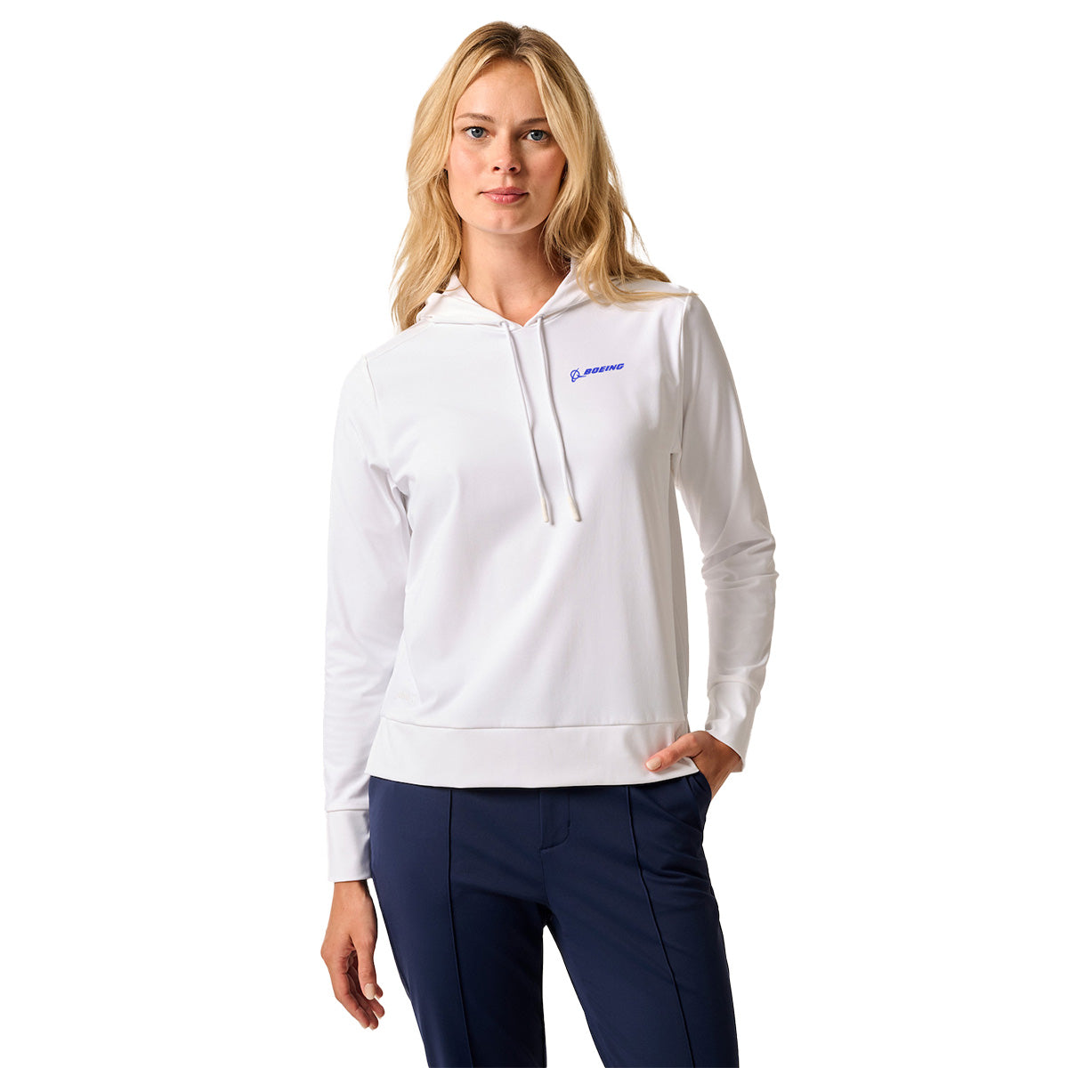 A woman with long blonde hair wears the johnnie-O Boeing Women’s Nalla Performance Hoodie in white, featuring a small blue BOEING logo on the chest and dark blue pants, standing with one hand in her pocket against a plain background.