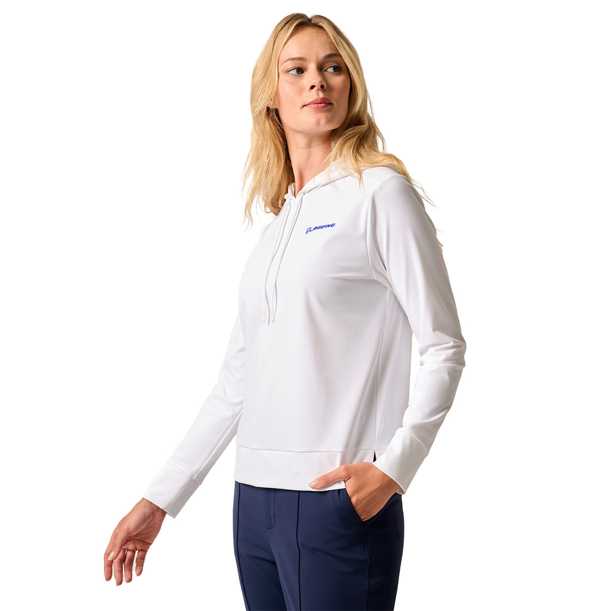 A woman wearing the johnnie-O Boeing Women’s Nalla Performance Hoodie in white, featuring moisture-wicking fabric and the BOEING logo, pairs it with navy blue pants and stands with one hand in her pocket against a white background.