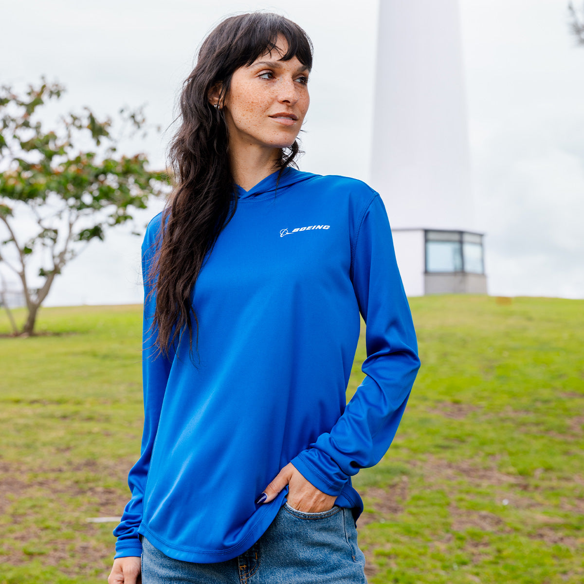 A woman with long dark hair stands outdoors on grass near a white lighthouse, wearing the BOEING Signature Logo Women's UV Hoodie in bright blue. She looks to the side with one hand in her jeans pocket; trees and a cloudy sky are behind her.
