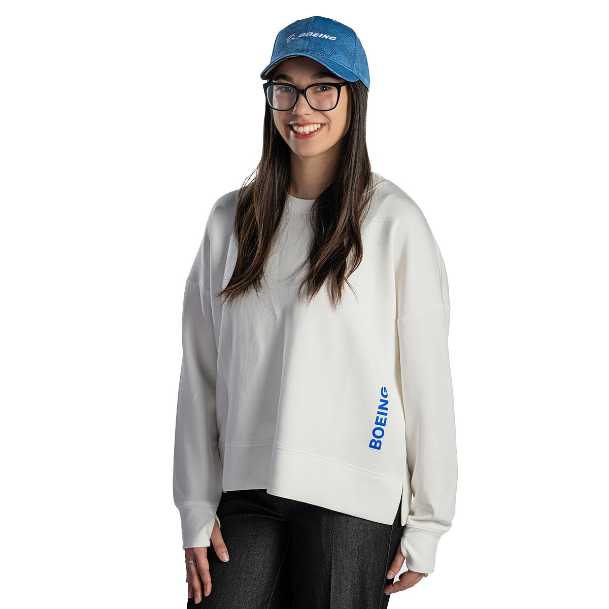 A woman with long brown hair and glasses smiles in a BOEING Women's Split-Hem Crewneck Sweatshirt and black pants, wearing a blue cap against a plain white background.