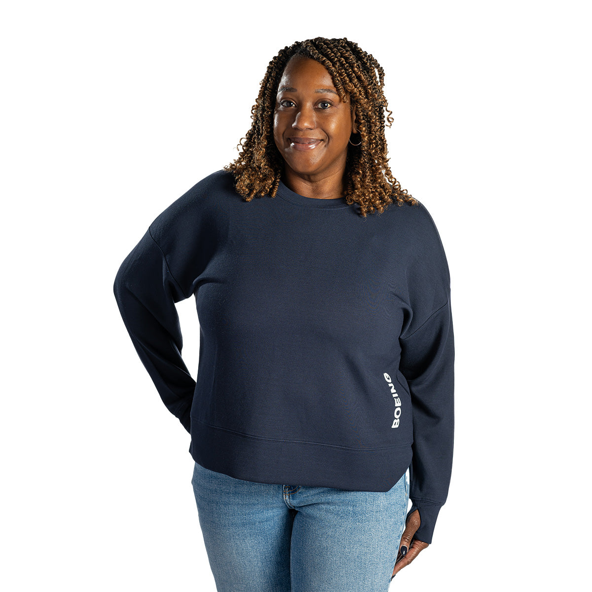 A woman with curly hair smiles while wearing the Boeing Women's Split-Hem Crewneck Sweatshirt by BOEING in navy blue, paired with light blue jeans, standing against a plain white background. "Boeing" is printed near the sweatshirt's hem.