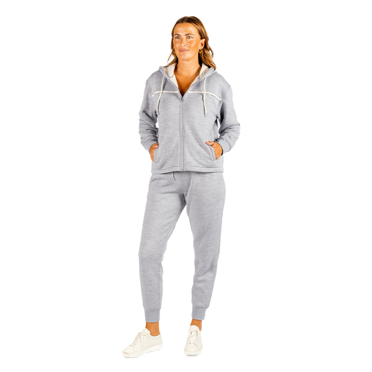 A woman wears a light gray zip-up hoodie with a cream-colored hood lining and Boeing Women’s Luxe Plush Jogger pants, paired with white sneakers.