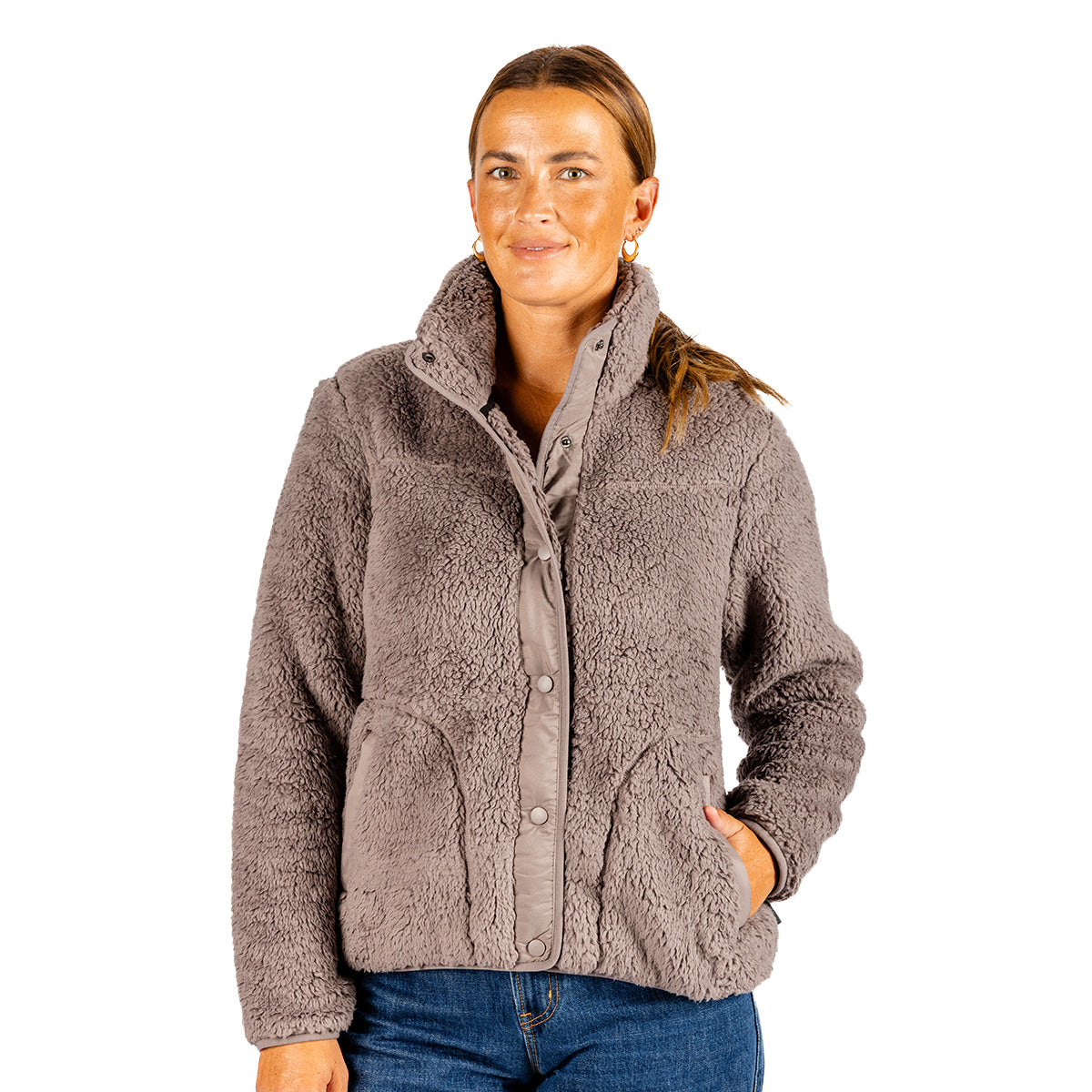 A woman wears a taupe Boeing Women’s Sierra Sherpa Pile Jacket, featuring a high-collar, polyester sherpa fabric, a snap-button front, and side pockets.