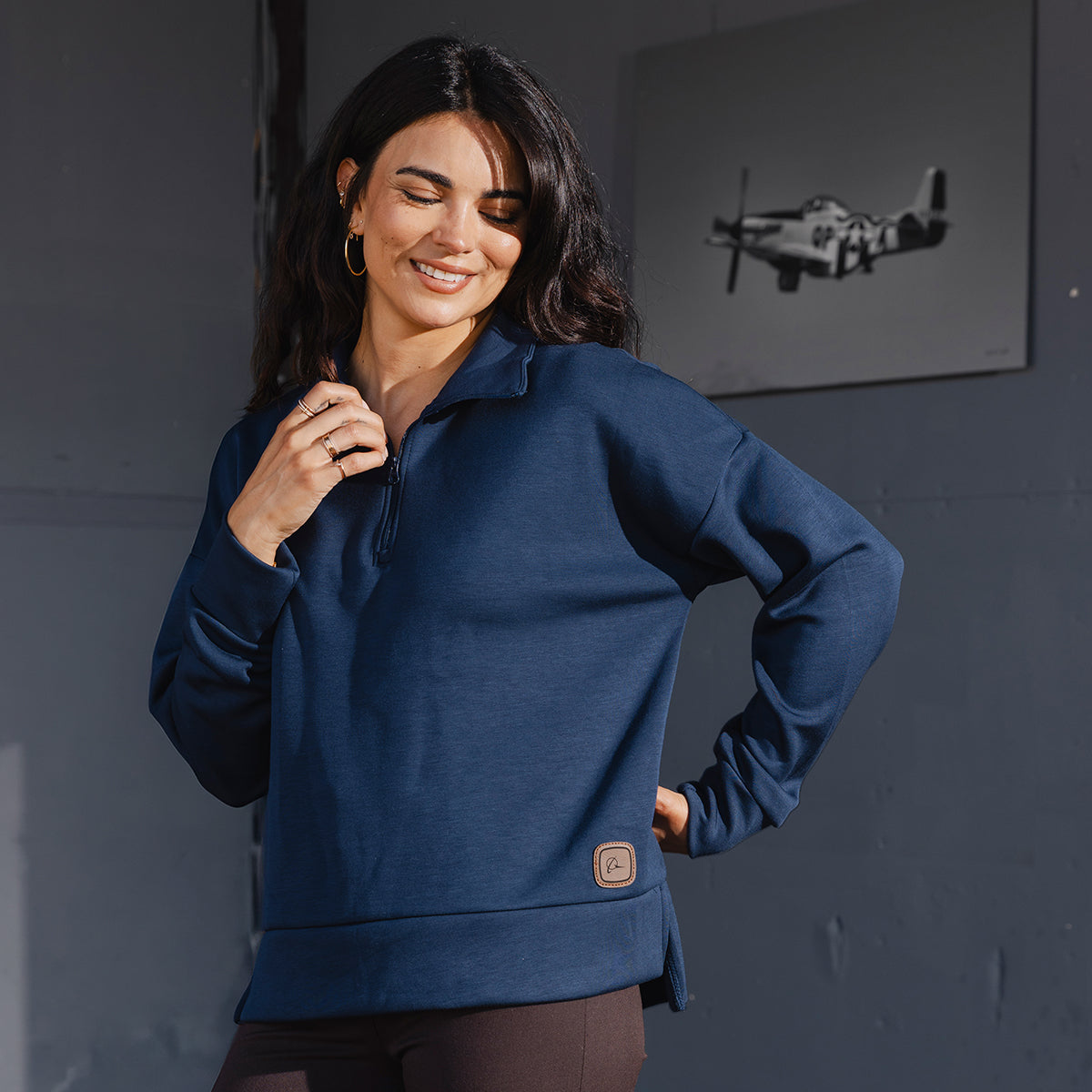 A woman wearing the Boeing Symbol Patch Women's Quarter-Zip by BOEING stands before a gray wall adorned with a black-and-white airplane photo, reflecting aerospace heritage. She smiles with her eyes closed, hand on her hip, accessorized with hoop earrings and dark pants.