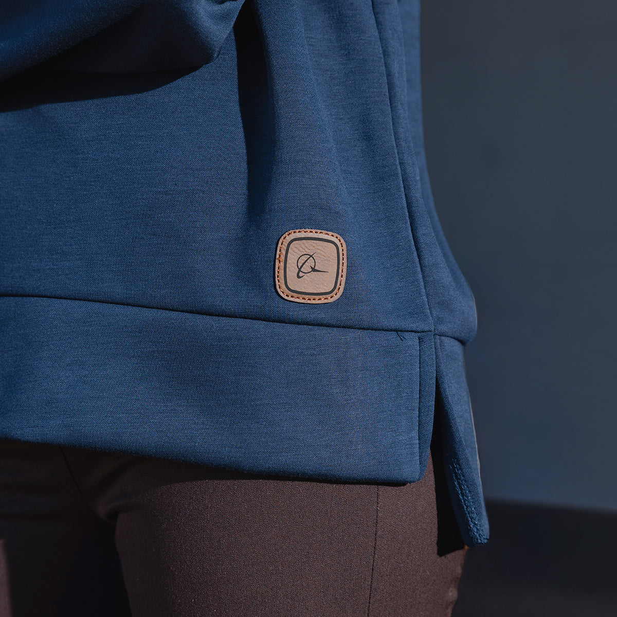 Close-up of a person wearing the Boeing Symbol Patch Women's Quarter-Zip by BOEING in blue, featuring a small brown square logo patch near the hem and paired with dark pants—a subtle nod to aerospace heritage.