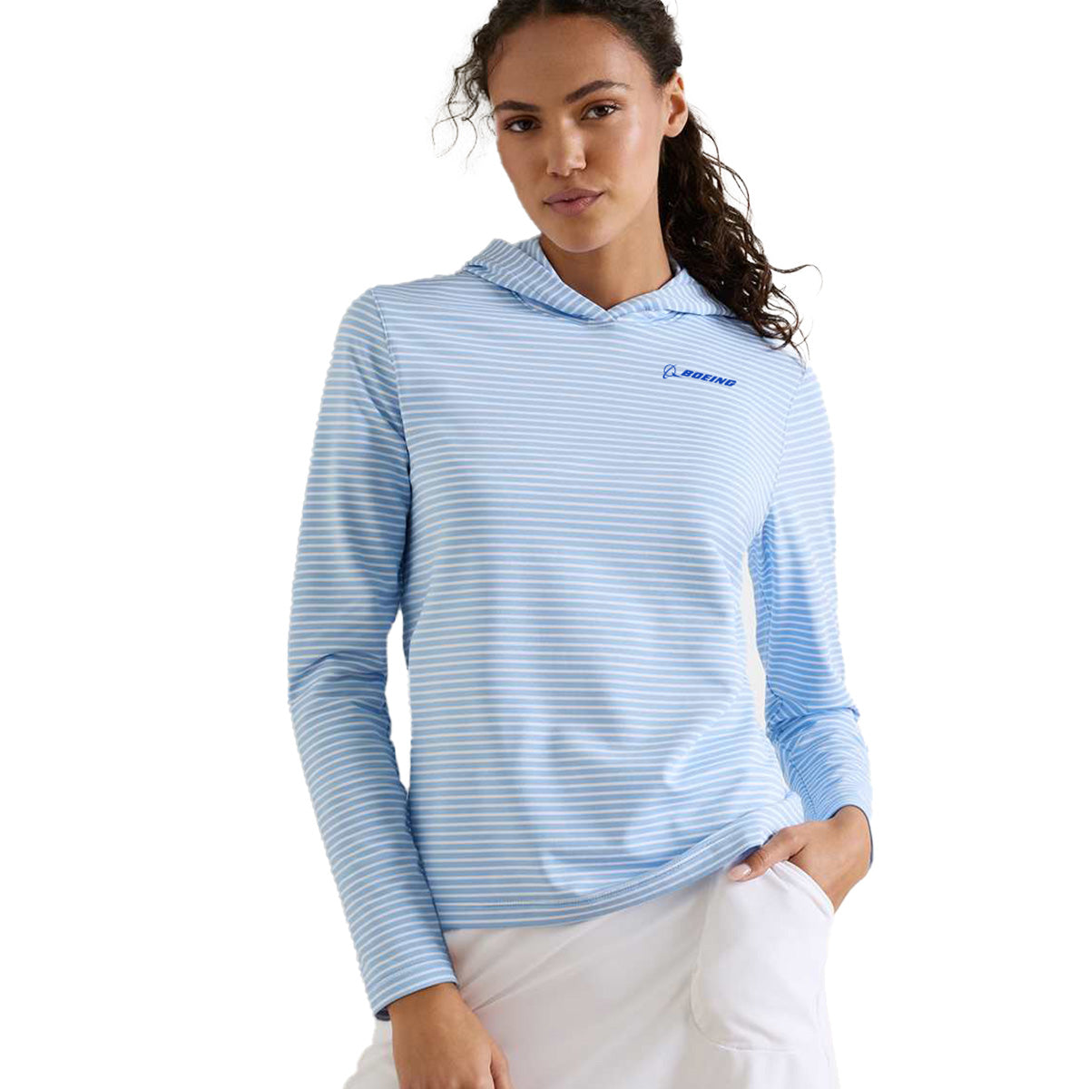 A woman stands against a plain background with one hand in the pocket of her white skirt, wearing the Peter Millar Boeing Women’s Harbour Stripe Hoodie—a light blue and white striped long-sleeve hooded shirt featuring the Boeing logo and crafted from soft, moisture-wicking fabric.