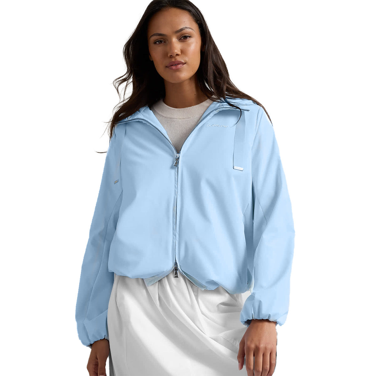 A woman stands against a plain white background wearing the Peter Millar Boeing Women’s Robinson Air Hooded Jacket in light blue—wind and water resistant with the BOEING logo—layered over a white top and skirt.