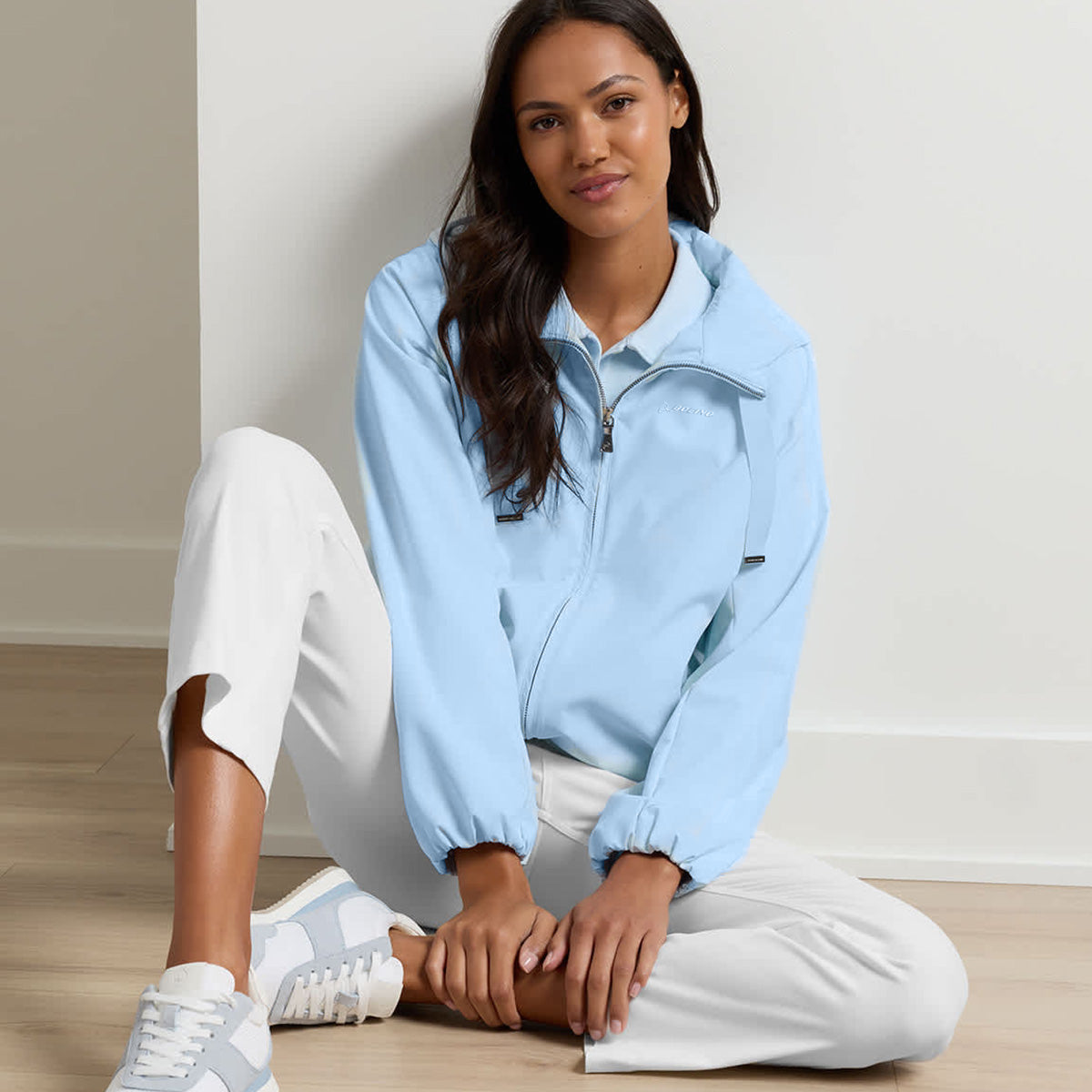 A woman with long, dark hair and a relaxed expression sits on a light wooden floor against a white wall, wearing the Peter Millar Boeing Women’s Robinson Air Hooded Jacket in light blue by BOEING. She has paired it with white pants and blue-and-white sneakers.