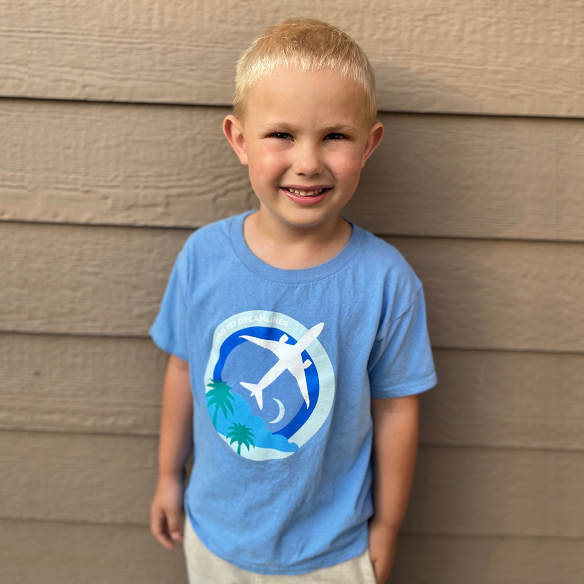 A young blonde child smiles in front of a wooden wall, wearing the Boeing 787 Dreamliner Skyward Kids T-Shirt by Boeing, featuring aviation graphics with an airplane and palm trees.