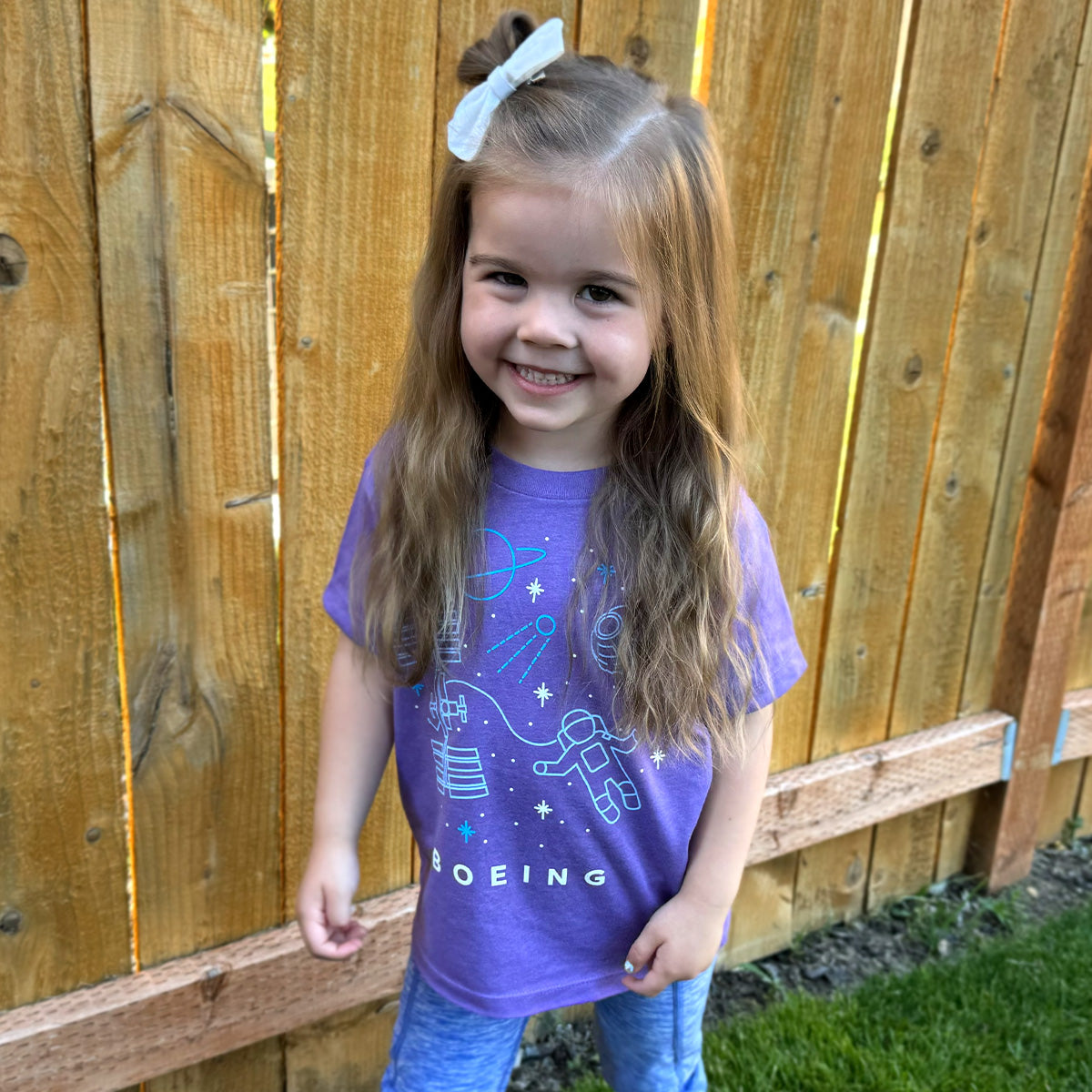A young girl with long hair stands smiling by a wooden fence, wearing a Boeing Space Doodle Toddler T-Shirt paired with blue pants.