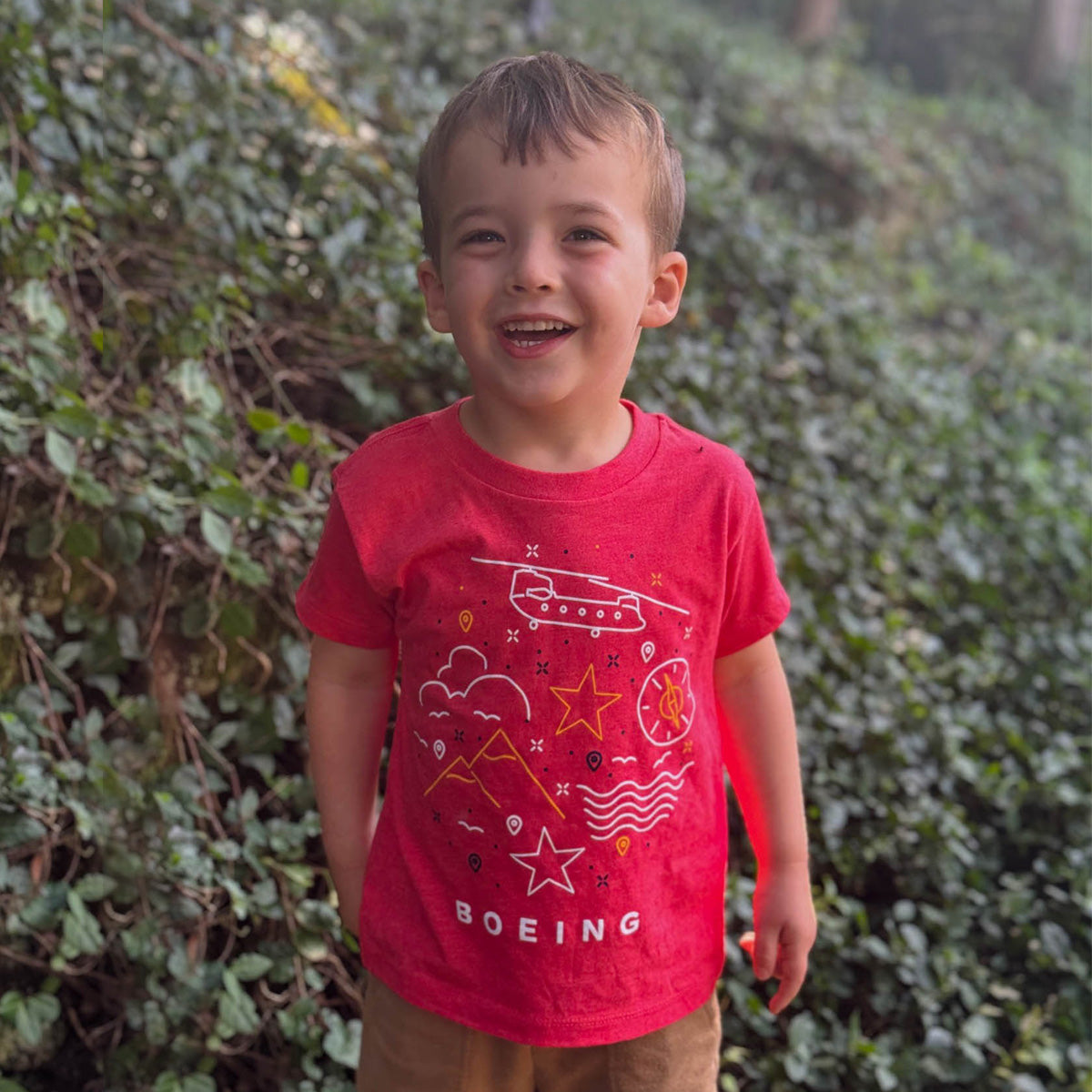 A young boy smiles outdoors in a vibrant green setting, wearing a Boeing Defense Doodle Toddler T-Shirt featuring a plane from Boeing Defense, stars, and the word BOEING.
