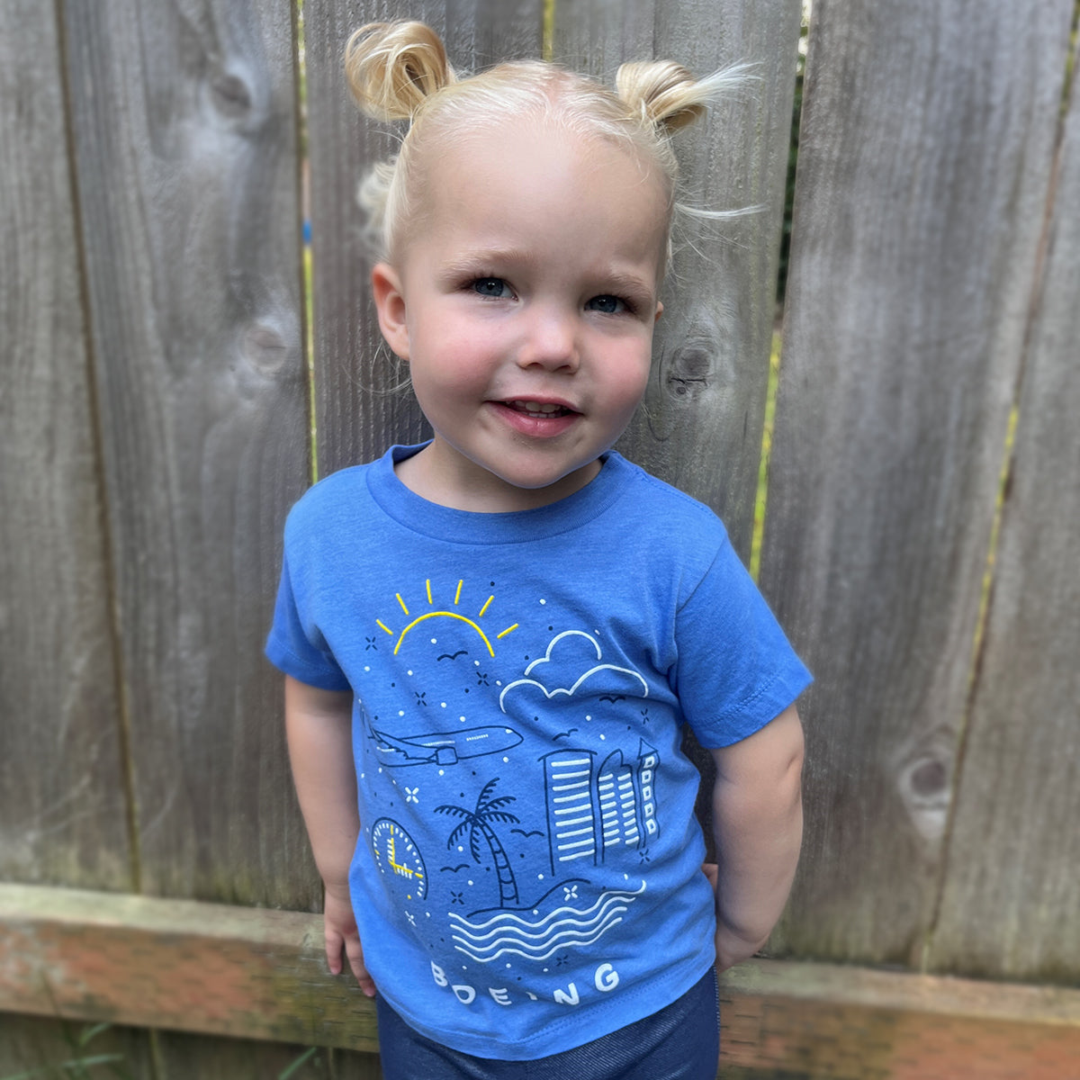 A smiling young child with blond hair styled in buns stands before a wooden fence, wearing a blue Boeing Commercial Doodle Toddler T-Shirt featuring a screen-printed graphic of the sun, clouds, palm trees, and buildings.