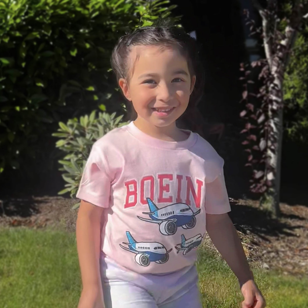 A young girl with braided hair stands on the grass, wearing a pink Boeing Toddler Varsity Pudgy T-Shirt featuring cartoon airplanes. Her smile is as bright as the lush greenery, and her Airlume cotton shirt ensures shes both stylish and comfortable.