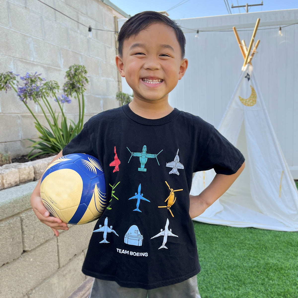 A smiling child stands in a backyard with a soccer ball, wearing the Boeing Kids Team Boeing T-Shirt, a classic fit made of 100% ring spun cotton featuring aircraft illustrations. A small white teepee peeks through the greenery behind them.