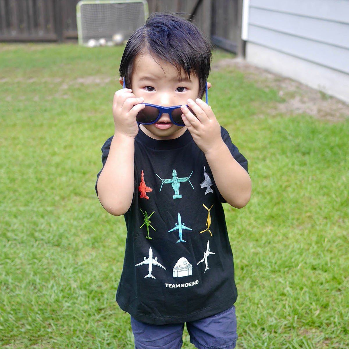 A child proudly wears a Boeing Toddler Team Boeing T-Shirt, made from soft Airlume combed and ring-spun cotton, on the lawn. Theyre holding blue sunglasses up to their face with a soccer goal in the background.