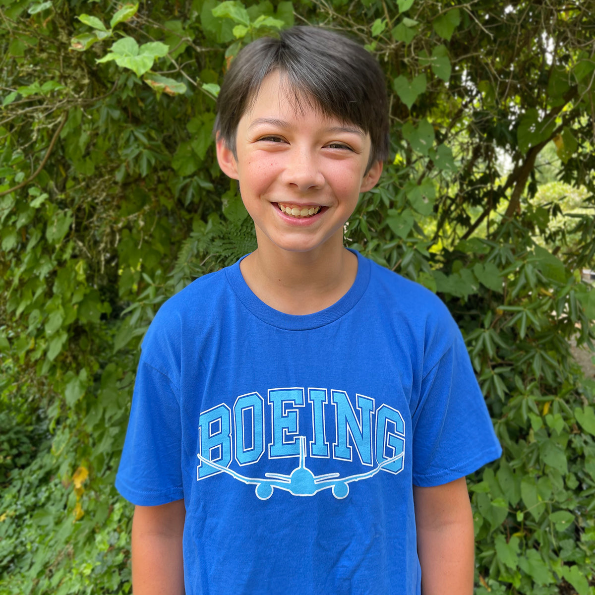 A young person beams in a Boeing Kids Varsity Graphic T-Shirt, featuring a classic fit blue design with BOEING and an airplane graphic. Made from 100% ring-spun cotton, the shirt pops against lush green foliage.