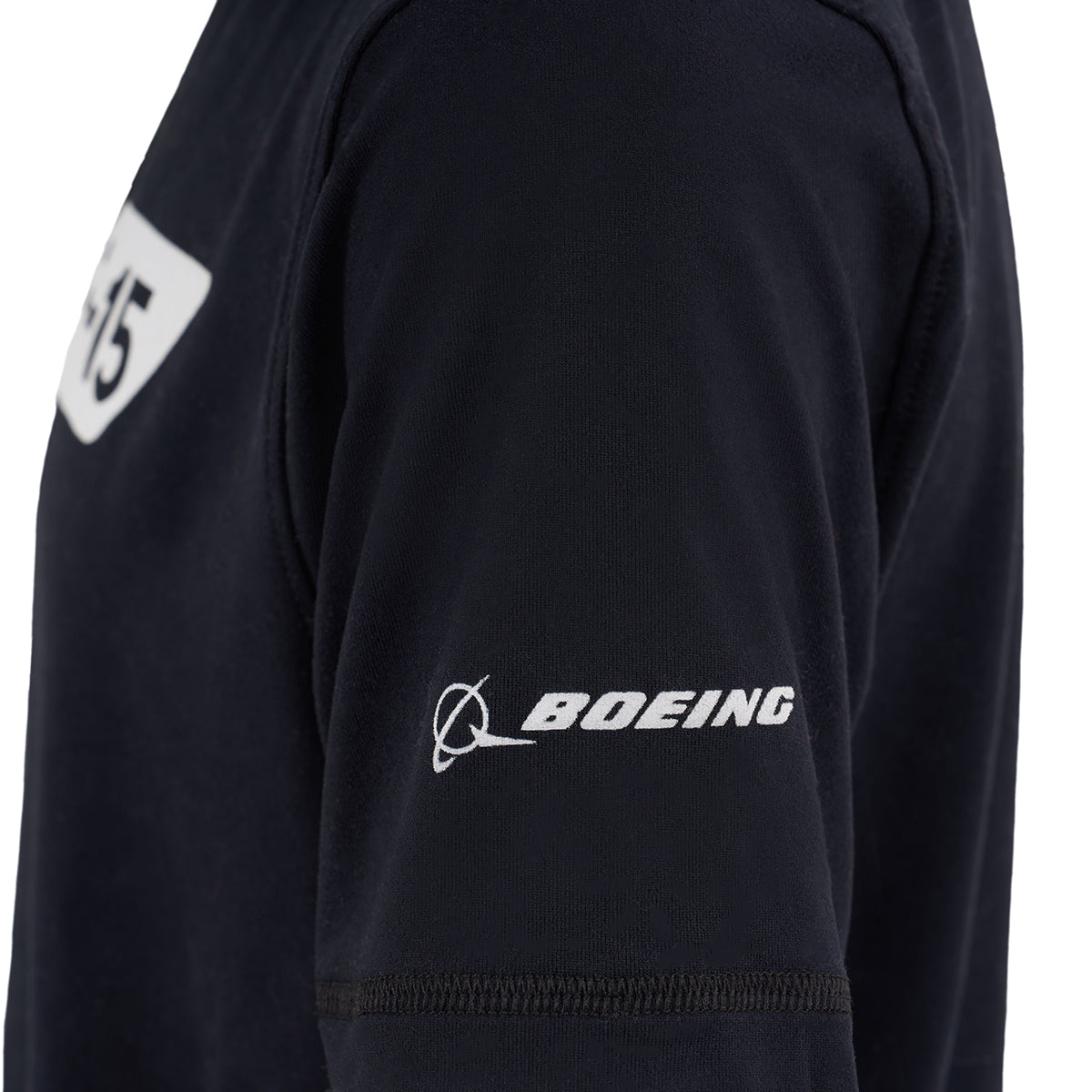 A close-up of a child’s upper arm in a black Boeing F-15 Eagle Velocity Kids’ Tech Stretch T-Shirt, featuring a white BOEING logo embroidered on the sleeve and a partially visible white “45” patch on the chest.