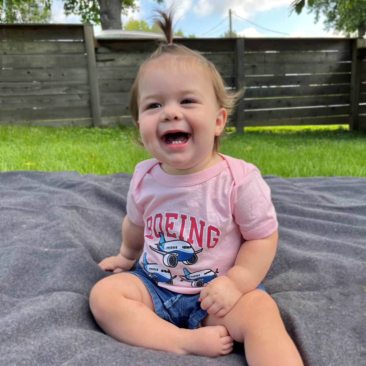 A smiling toddler with a tiny ponytail sits on a gray blanket in the backyard, wearing a Boeing Baby Varsity Pudgy Onesie with airplane graphics. Denim shorts complete the ensemble, while a wooden fence frames the lush green grass behind.